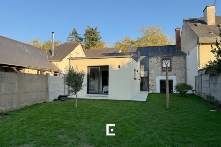 Maison mêlant architecture traditionnelle en pierre et extension moderne beige avec grandes baies vitrées, toiture plate et jardin clôturé, illustrant une intégration harmonieuse entre ancien et contemporain