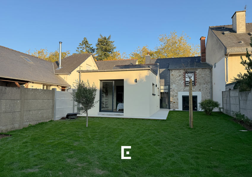 Maison mêlant architecture traditionnelle en pierre et extension moderne beige avec grandes baies vitrées, toiture plate et jardin clôturé, illustrant une intégration harmonieuse entre ancien et contemporain