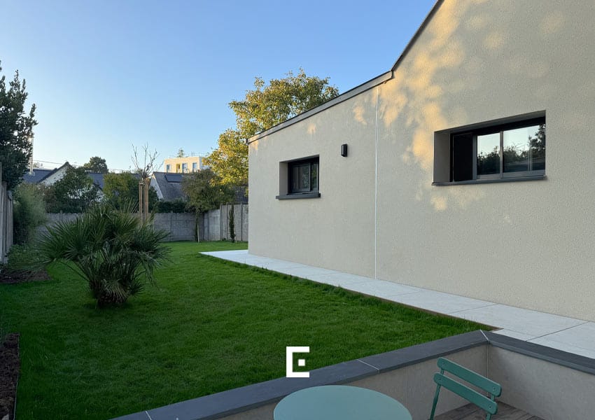 Maison contemporaine aux murs crème avec une petite fenêtre, vue depuis une terrasse carrelée équipée d’une table et de chaises, donnant sur une pelouse verte parfaitement entretenue