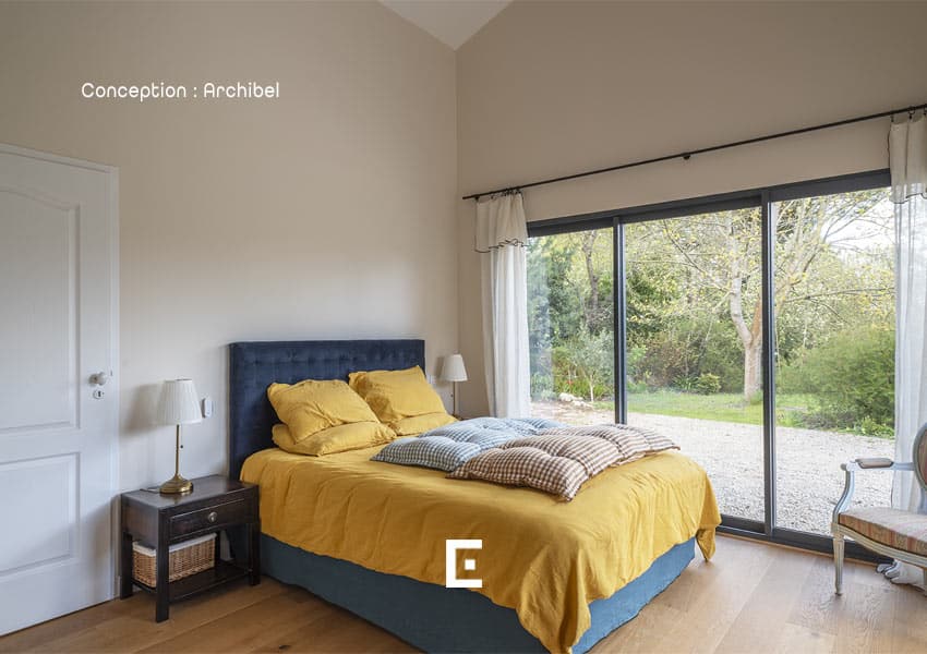 Chambre chaleureuse avec un lit à tête de lit bleu, linge de lit jaune, deux tables de chevet et une grande baie vitrée donnant sur un jardin arboré, laissant entrer une lumière douce et naturelle.