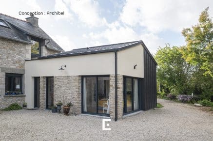 Vue d’une extension moderne avec façades en enduit clair, pierre et bardage sombre, comportant de larges baies vitrées et reliée à une maison en pierre traditionnelle, le tout entouré d’une cour en gravier et de végétation.