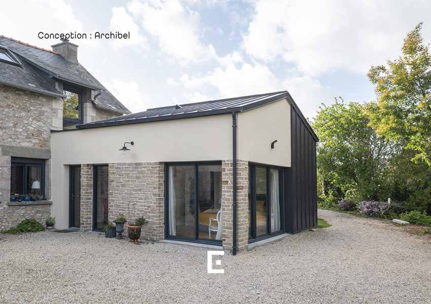 Vue d’une extension moderne avec façades en enduit clair, pierre et bardage sombre, comportant de larges baies vitrées et reliée à une maison en pierre traditionnelle, le tout entouré d’une cour en gravier et de végétation.