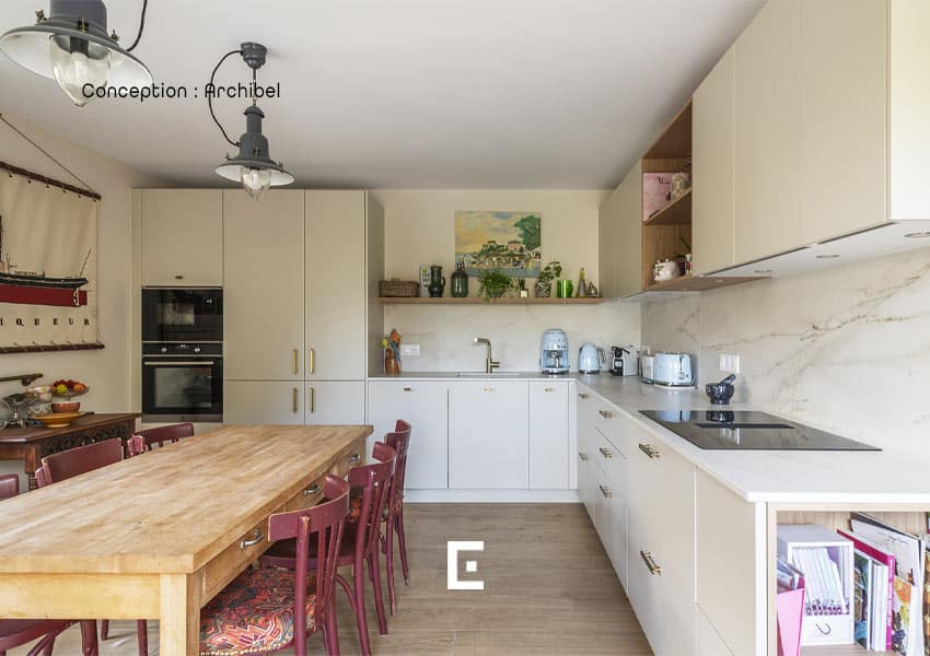 Cuisine lumineuse avec meubles clairs, grand plan de travail en L, table en bois entourée de chaises rouges, électroménager pastel sur le comptoir et décorations murales, dans un espace au style chaleureux et soigné.