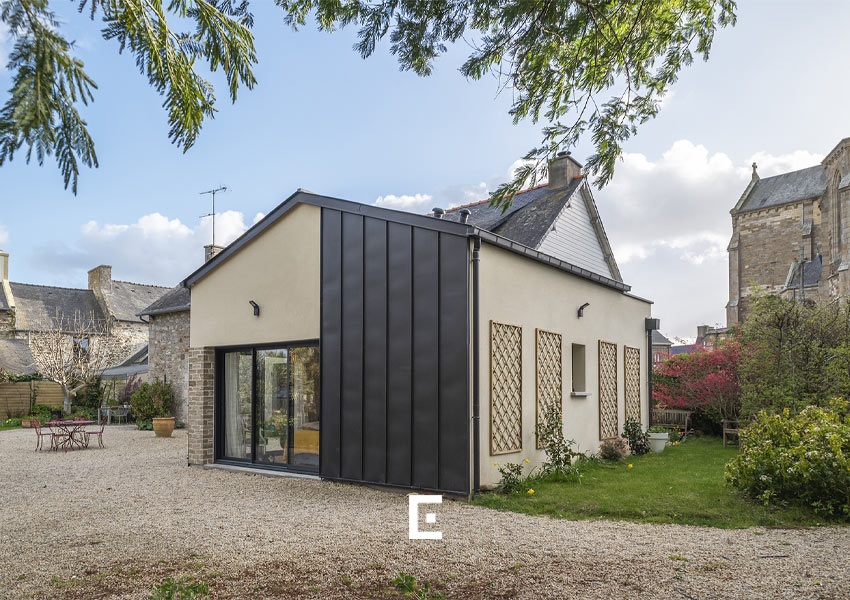Extension contemporaine avec bardage métallique sombre et enduit clair, accolée à une maison en pierre, donnant sur une cour en gravier avec mobilier extérieur et entourée de jardin et de végétation sous un ciel partiellement nuageux.