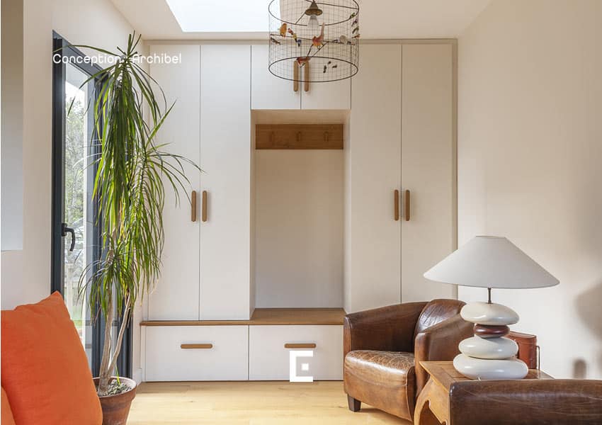 Entrée lumineuse avec un ensemble de placards blancs dotés de poignées en bois, un espace central avec banc et patères, une plante haute près d’une baie vitrée, ainsi qu’un fauteuil en cuir et une lampe design posés sur un meuble en bois.