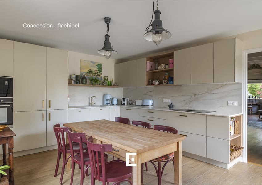 Cuisine lumineuse dotée de meubles clairs et d’un long plan de travail en marbre clair, comprenant une grande table en bois entourée de chaises rouges, des étagères ouvertes avec objets décoratifs, ainsi que des suspensions industrielles au plafond.