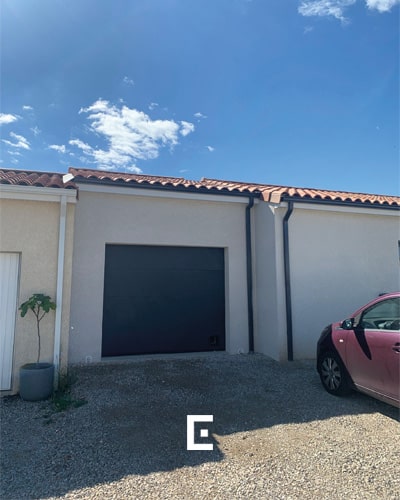 Zoom sur une porte de garage électrique noire intégrée à une extension d’une maison blanche de plain-pied.