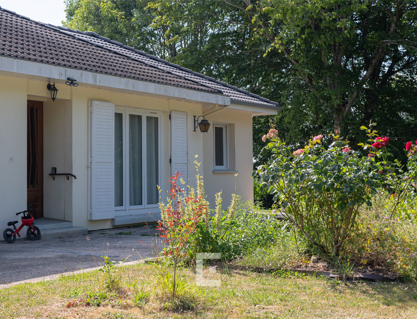 Façade d'une maison de plain-pied avec extension maçonnée beige, volets battants blancs et massif de rosiers dans le jardin.