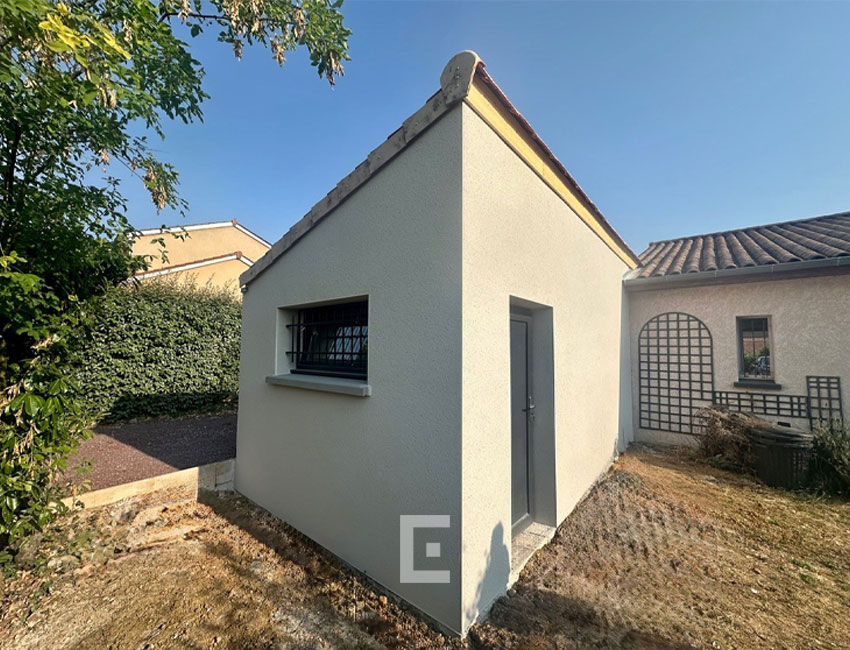 Extension d’une maison individuelle en maçonnerie avec façade beige claire, petite fenêtre à barreaux et porte vitrée anthracite sous un toit en tuiles.