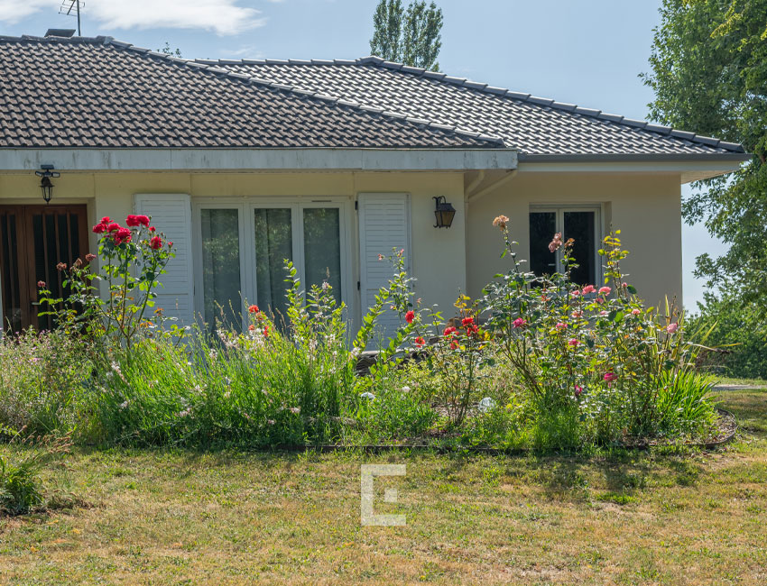 Vue de face d'une maison beige avec toiture en tuiles foncées et parterre de fleurs au premier plan.