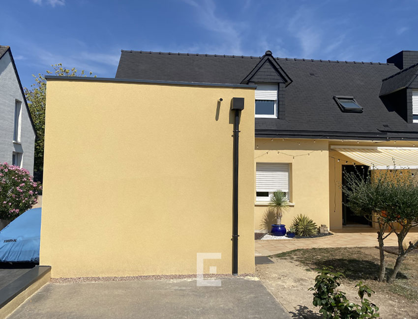 Extension de maison à façade jaune avec toiture plate, accolée à une maison existante avec toit en ardoise.
